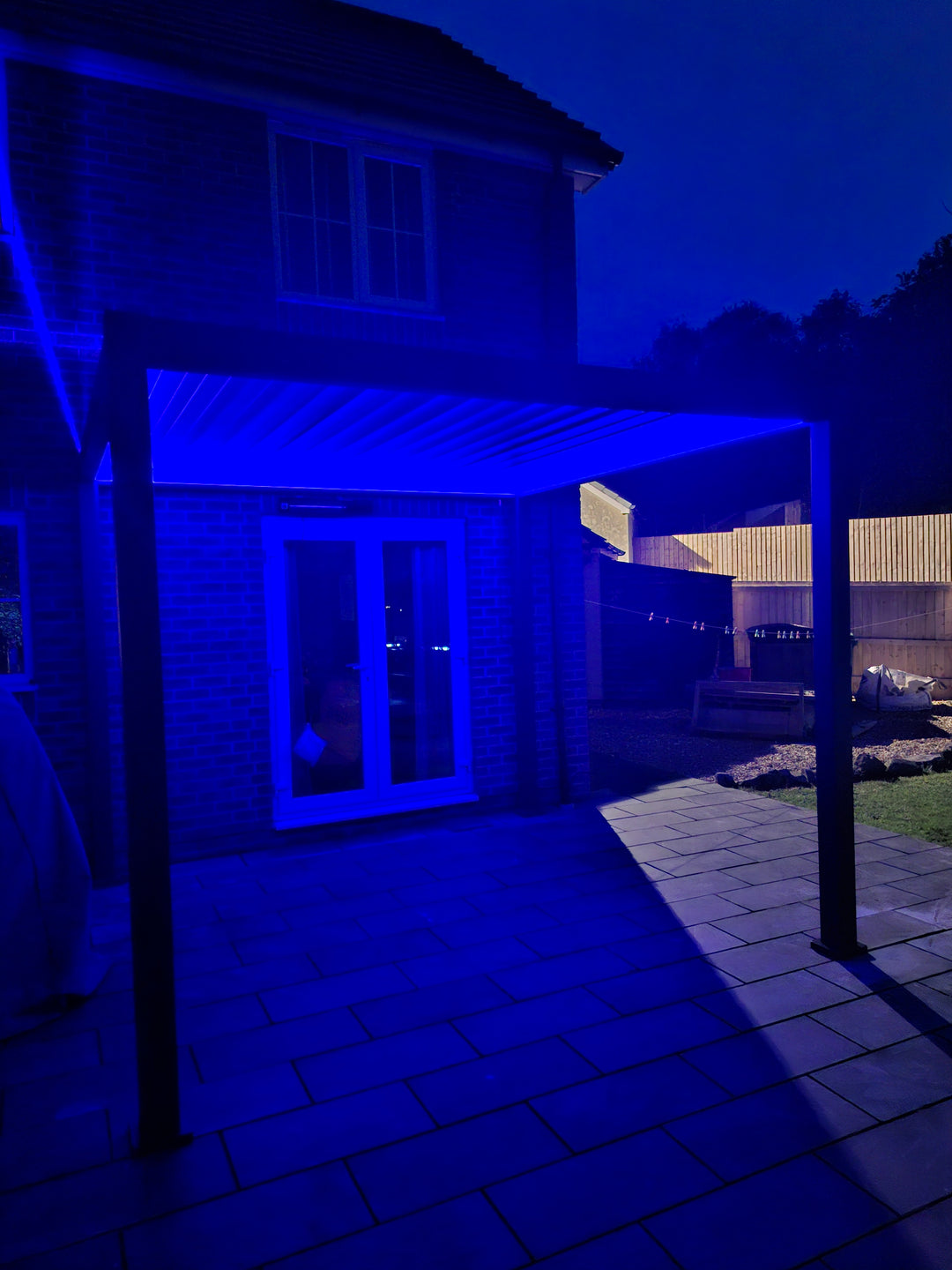 Nova Titan LED Pergola at night with dark blue LED lighting under the louvers on a patio next to a house with doors opening to the Pergola.