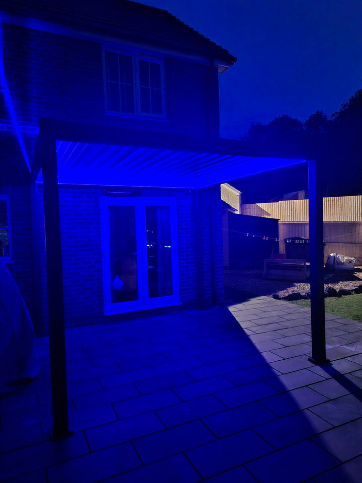 Nova Titan LED Pergola at night with dark blue LED lighting under the louvers on a patio next to a house with doors opening to the Pergola.