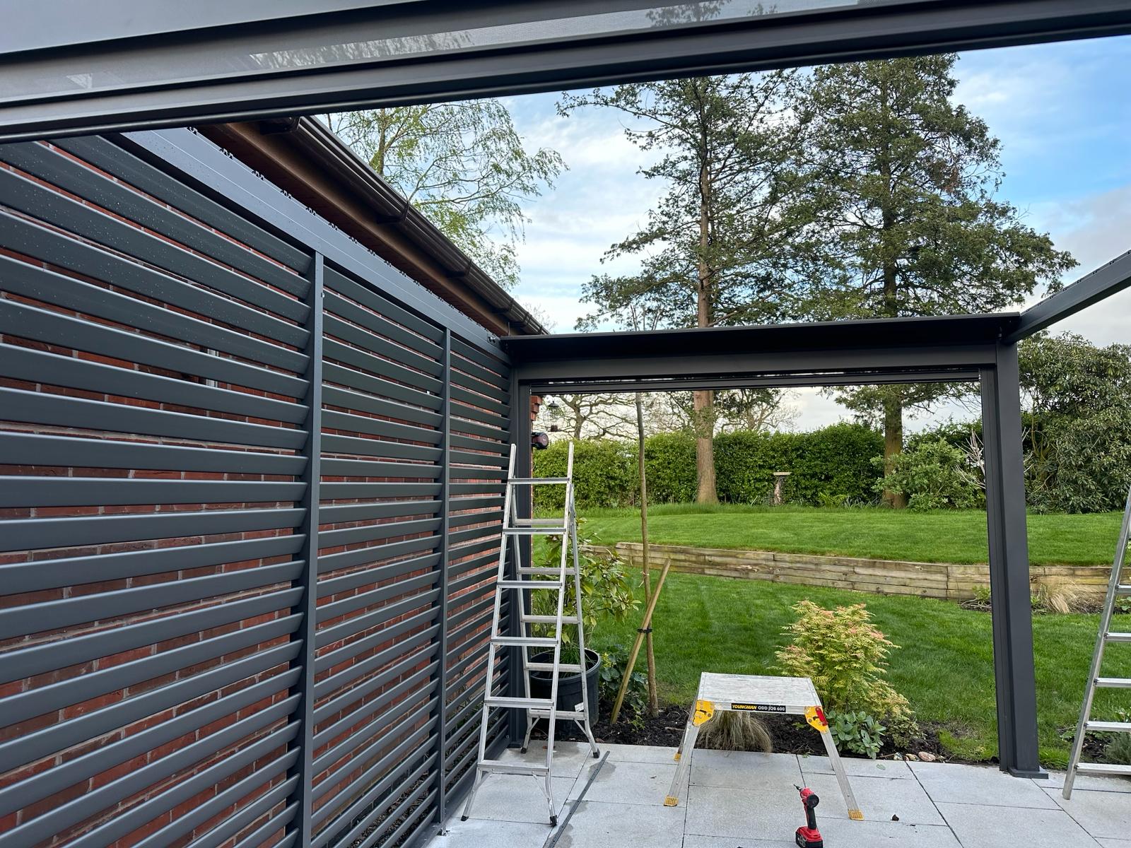 Grey Titan Pergola under construction with no roof louvers, Grey Pergola and side walls with garden to rear with large tree.