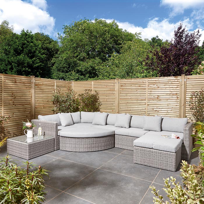 Rowlinson Marbella Multifunction Rattan Garden Furniture, with grey rattan and light grey cushions with a fence behind and bush at the back. Corner section image with straight sofa at the side
