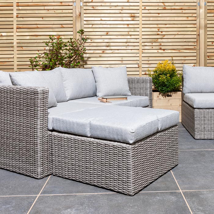 Rowlinson Marbella Multifunction Rattan Garden Furniture, with grey rattan and light grey cushions with a fence behind and bush at the back. 2 seat sofa and foot stool