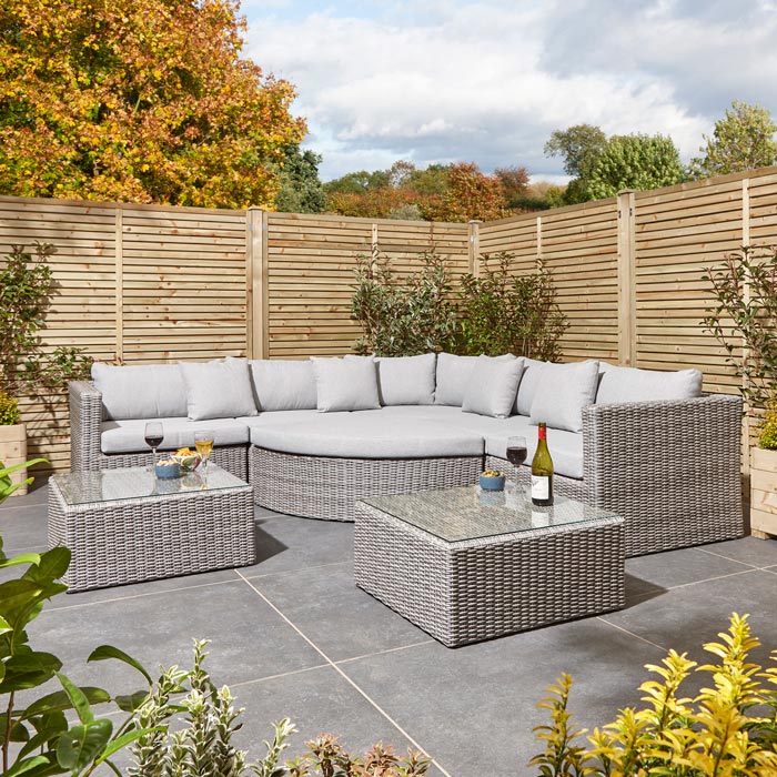 Rowlinson Marbella Multifunction Rattan Garden Furniture, with grey rattan and light grey cushions with a fence behind and bush at the back. Corner section image with two coffee tables.