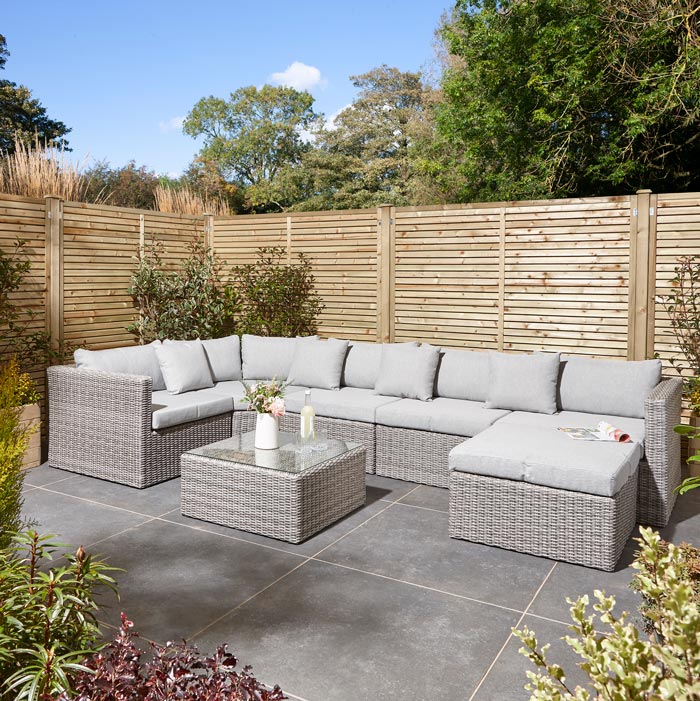 Rowlinson Marbella Multifunction Rattan Garden Furniture, with grey rattan and light grey cushions with a fence behind and bush at the back. Long sofa set section with coffee table.
