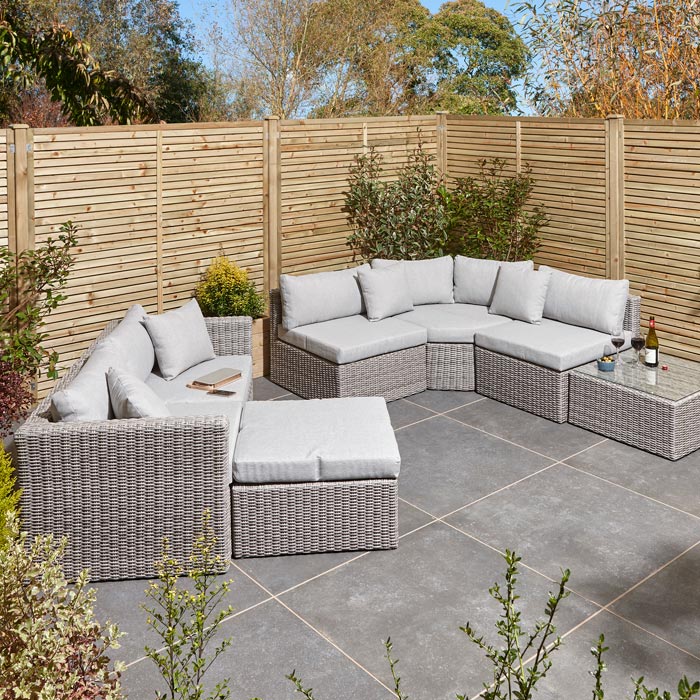 Rowlinson Marbella Multifunction Rattan Garden Furniture, with grey rattan and light grey cushions with a fence behind and bush at the back. Corner section image and 2 seater sofa the opposite side.