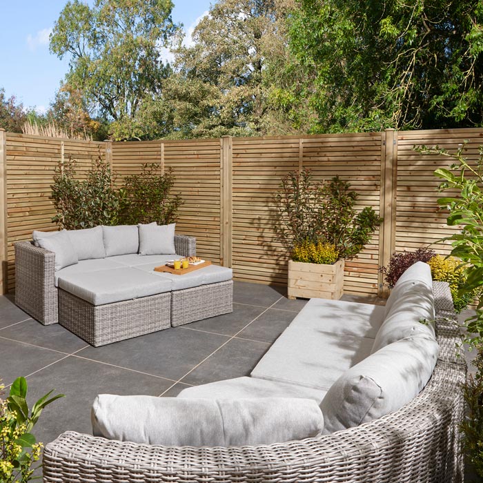 Rowlinson Marbella Multifunction Rattan Garden Furniture, with grey rattan and light grey cushions with a fence behind and bush at the back. Corner section image and 2 seater sofa with foot stools.