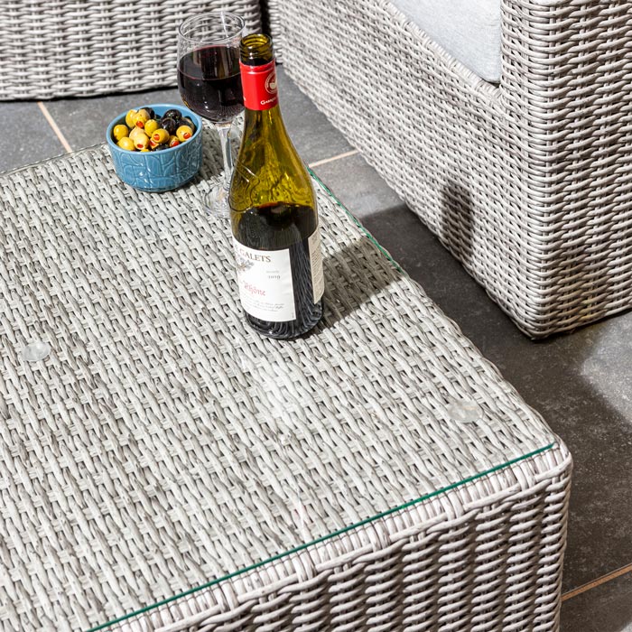 Rowlinson Marbella Multifunction Rattan Garden Furniture, with grey rattan and light grey cushions with a fence behind and bush at the back. Close up of coffee table with glass of wine