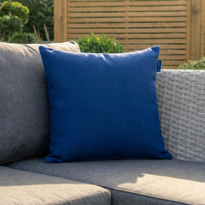 A royal blue square throw pillow resting on a gray outdoor patio sofa next to a woven wicker chair, with a wooden slat fence and greenery in the background. Alba Scatter cushion NOVA Whitestores from Midland Garden Furniture.