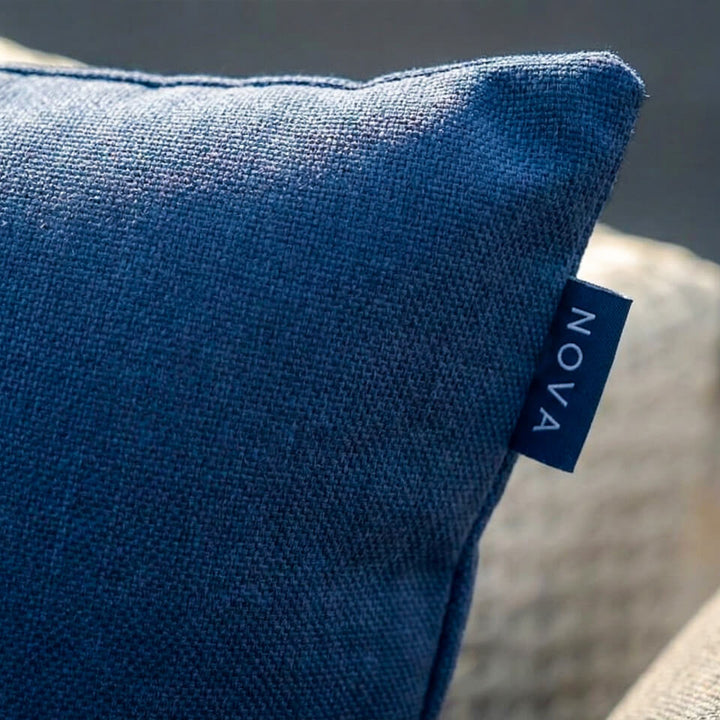 A close-up of a textured blue throw pillow with a small dark blue fabric tag on the side that reads 'NOVA' in white text, resting on a light beige woven chair.  Alba Scatter cushion NOVA Whitestores from Midland Garden Furniture.