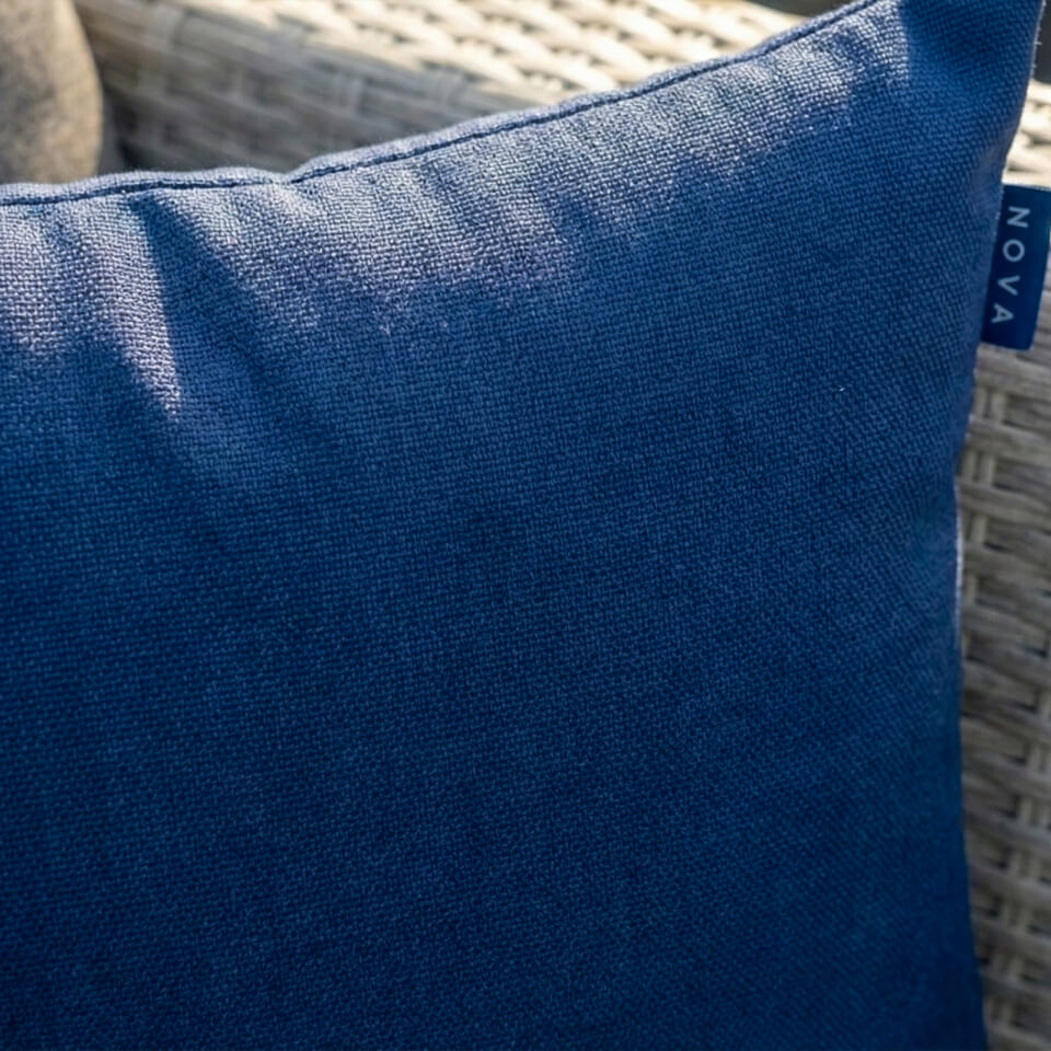 A close-up of a blue textured outdoor throw pillow with a visible 'NOVA' brand tag, resting on a woven wicker patio chair. Alba Scatter cushion NOVA Whitestores from Midland Garden Furniture.