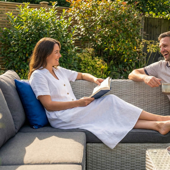 A woman in a white dress sits on a gray outdoor sectional sofa reading a book, with a blue pillow beside her and a man partially visible on the right in a garden setting. A blue textured outdoor cushion with a visible 'NOVA' brand tag, resting on a woven wicker patio chair. Alba Scatter cushion NOVA Whitestores from Midland Garden Furniture.