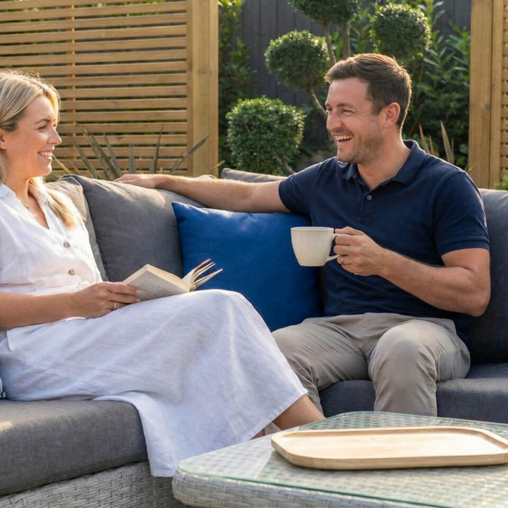 A man and a woman relax on a gray outdoor patio sofa, smiling and chatting. The man holds a white mug, the woman reads a book, and a glass coffee table with a wooden tray sits in the foreground. Alba Scatter cushion NOVA Whitestores from Midland Garden Furniture.
